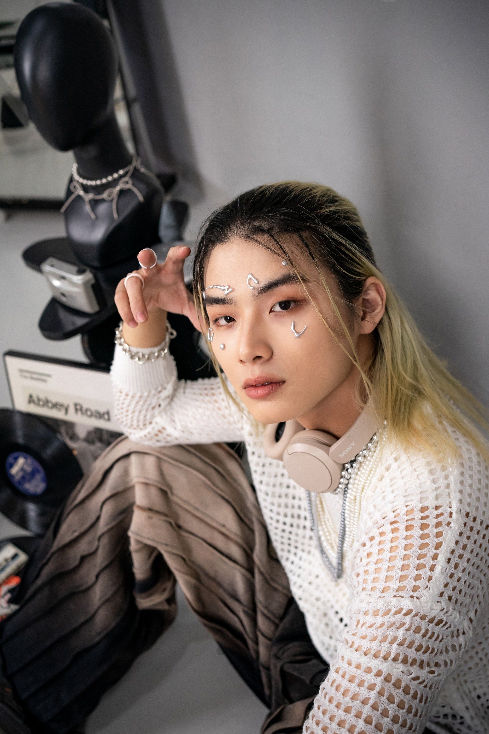 白い透かし編みのニットを着たTaichanのアップ。首元にヘッドホンと重ね付けのネックレス、顔にシルバーの装飾を着け、片手を顔の横に上げてカメラを見つめています。
